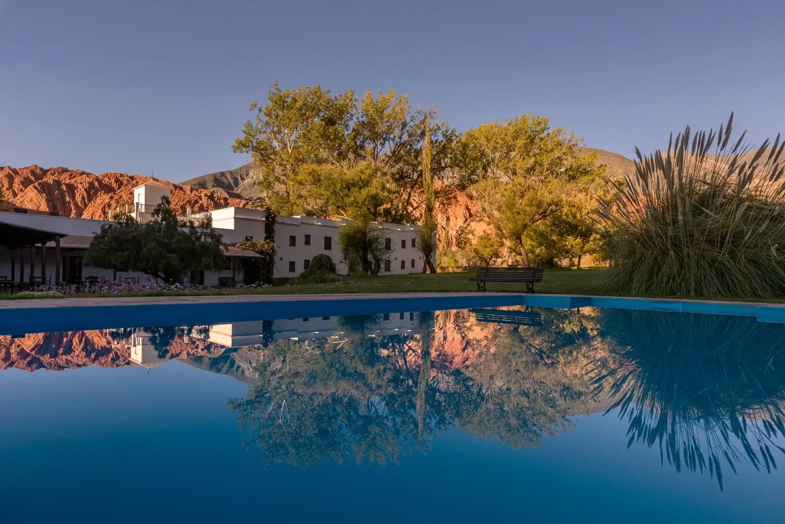 La pileta al atardecer reflejando los álamos, la casona blanca y los cerros rojizos