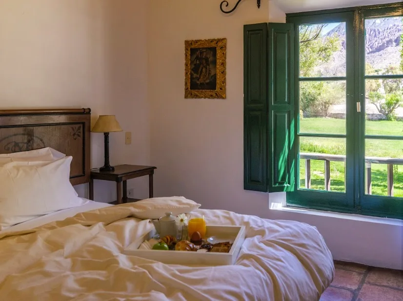 Cama tendida con bandeja de desayuno frente a una ventana de postigos verdes abierta al parque